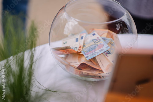 A glass bowl filled with euro banknotes sits on a white tablecloth, possibly used for donations, tips, or fundraising purposes.. photo