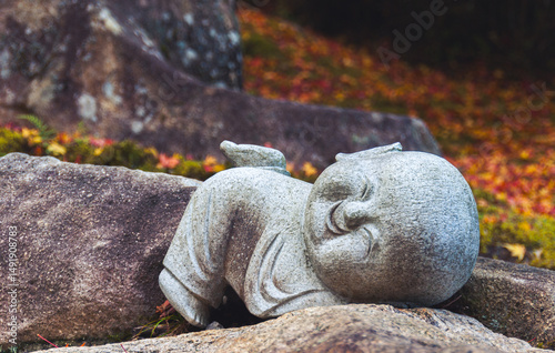 Smiling 'jizo' statue lies on the ground against a background of rock and autumn leaves at 'Daisho-in' temple
