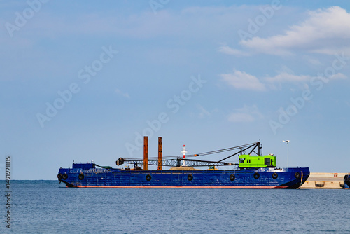A vibrant blue ship, equipped with cranes and various tools, is anchored close to the rocky coastline nearby