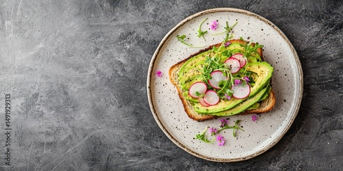 Avocado toast topped with radish, greens, and edible flowers served on a rustic plate over a textured gray background.