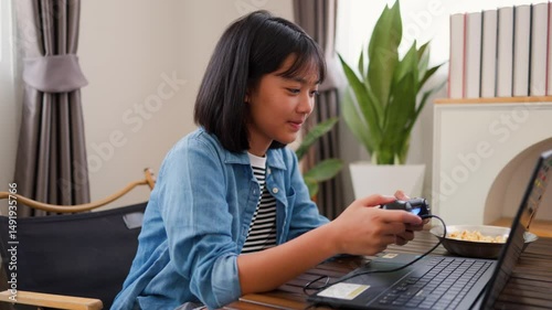Asian teenage girl using game controller while talking during online gaming session at home. Concept of digital communication, live streaming, and modern tech lifestyle for teens.