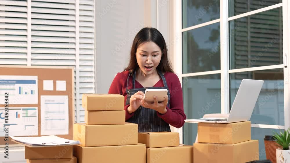 A stressed young female entrepreneur sits in her home office, frustrated and worried while managing online orders, packing boxes, and handling logistics.