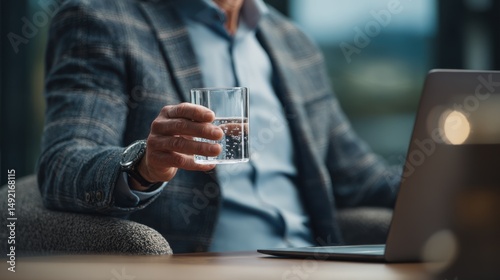Businessman holding a glass of water while working on a laptop in an office environment.