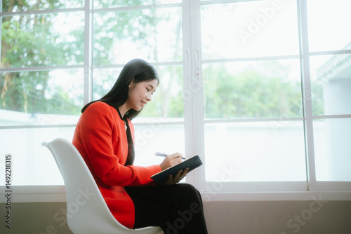 Free Download A young Asian businesswoman sits at her desk with a laptop, smiling confidently while working remotely on accounting and marketing tasks. - GFXLoad