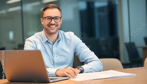 A young man with glasses smiling while using a smartphone, standing outdoors in a modern urban setting with glass buildings in the background. He is wearing a dark blue shirt.