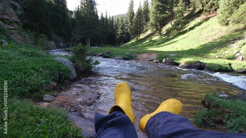 Legs in yellow EVA boots against the backdrop of a fast river, rocks and forest in the spring mountains.