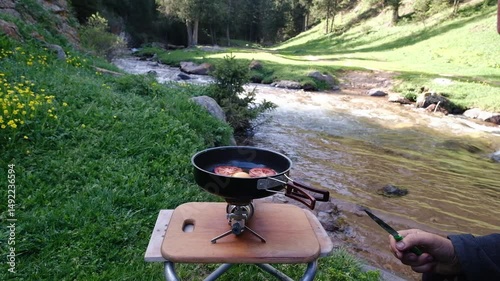 Hands of a man preparing scrambled eggs with tomatoes for breakfast on a portable gas stove, on a spring morning against the backdrop of a mountain river and forest. Cozy and authentic camping lifesty