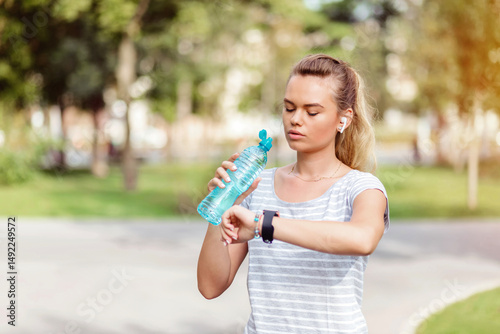 Young blonde woman jogging in park checking smartwatch and hydrating with water