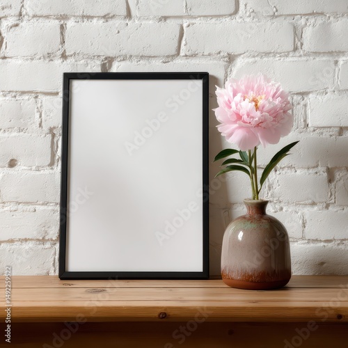 A large black frame stands empty beside a vase containing a pink flower. The setting features a rustic white brick wall, creating a serene ambiance
