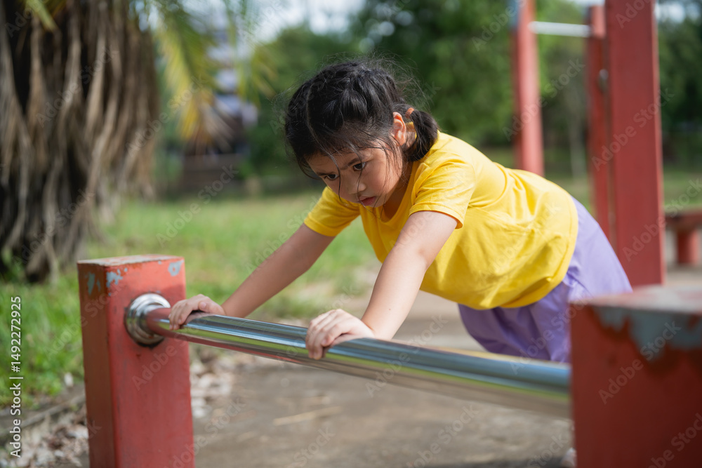 Obraz premium Asian Young girl exercising outdoors in a park, performing push-ups on a metal bar, demonstrating strength and determination in a vibrant yellow shirt