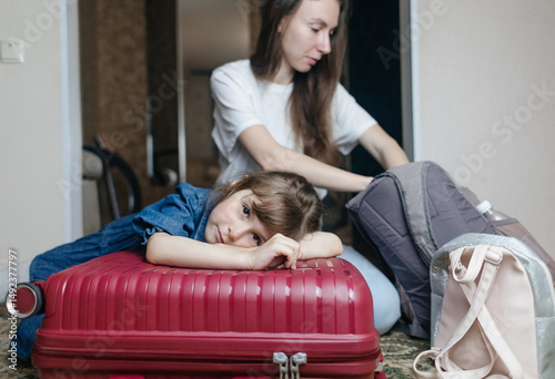 Tired 6 year girl lying on travel suitcase while mother is getting ready for trip. Cute child lost strength after packing bag and preparing for trip. Loving family, child care, home schooling concept