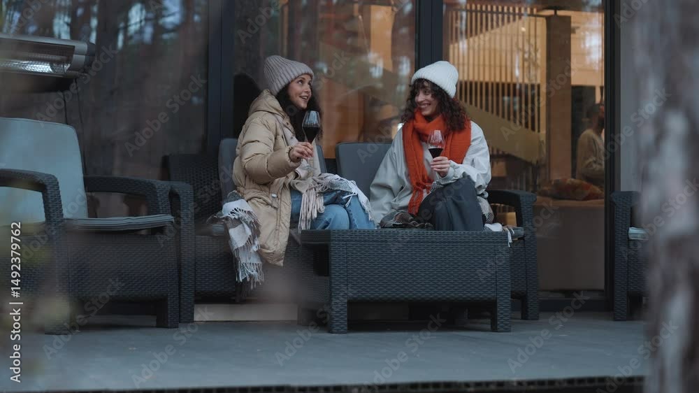 A European woman in her late 20s and a Latina woman in her 30s sitting on a sofa at a table with glasses of wine on a porch in front of a house with big windows and talking. dressed in beanies and jac A European woman in her late 20s and a Latina woman in her 30s sitting on a sofa at a table with glasses of wine on a porch in front of a house with big windows and talking. dressed in beanies and jac
