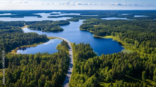 A road winds through green woods next to blue lakes in Finland during the summer, as seen from above.
