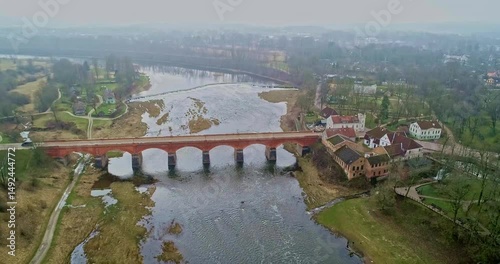 An aerial view shows a magnificent historic red brick bridge spanning a serene river Kuldiga Latvia