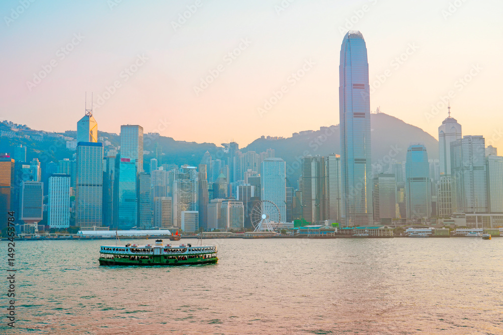 Fototapeta premium Classic green ferry at the pier on the route sails evening sunset view of tall glass skyscrapers in the financial district building, a symbol of the region's economic development. China, Hong Kong