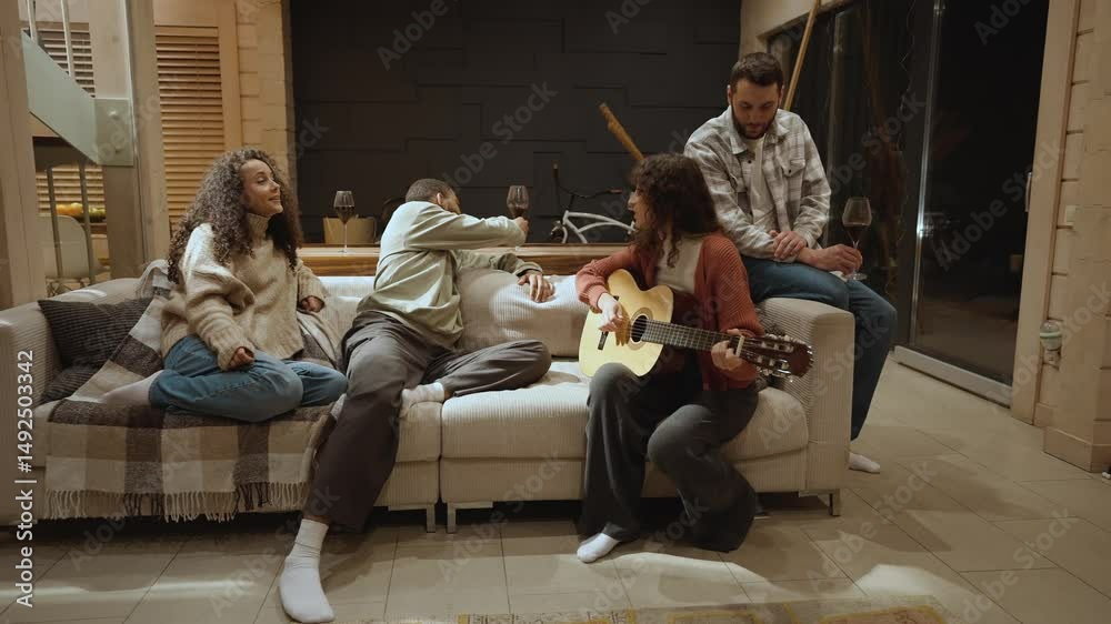 A multiethnic group of two men and one woman in their 30s sitting with glasses of wine on a white sofa around a White adult woman playing guitar and singing for them in a cozy living room