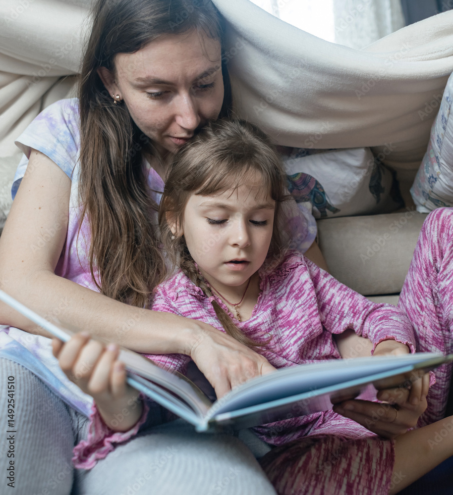 Mother and daughter sit in a hut made of a blanket and read a book. Pleasant time with children, quiet time before bed. Mother and daughter sit in a hut made of a blanket and read a book. Pleasant time with children, quiet time before bed.