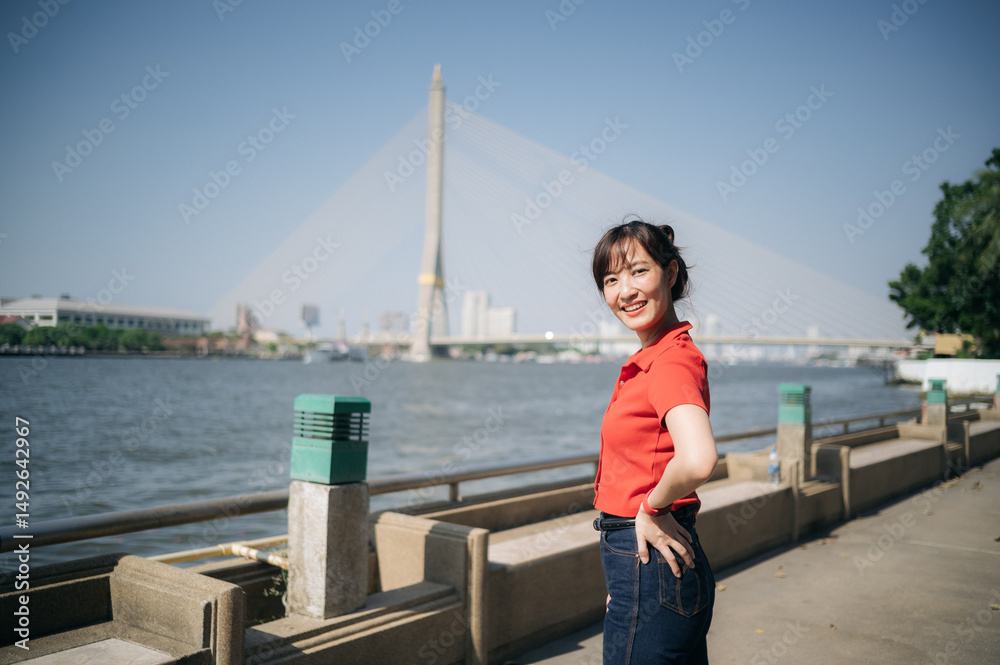 Obraz premium Southeast asian woman young female entrepreneur remote work small business owner businesswoman stands confidently by river with modern bridge in background under clear sky