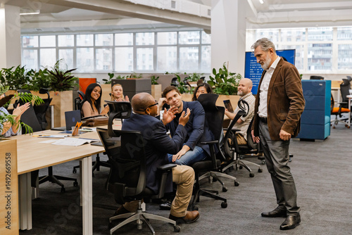 Фототапет The multiracial adult men and women interact and applaud during a business meeting in a modern open-plan office with desks, laptops, and windows in the background