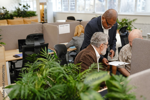 A Black bald man in a suit and eyeglasses is holding a tablet and showing it to two white-skinned men sitting on each side of him in a modern office with green plants.
