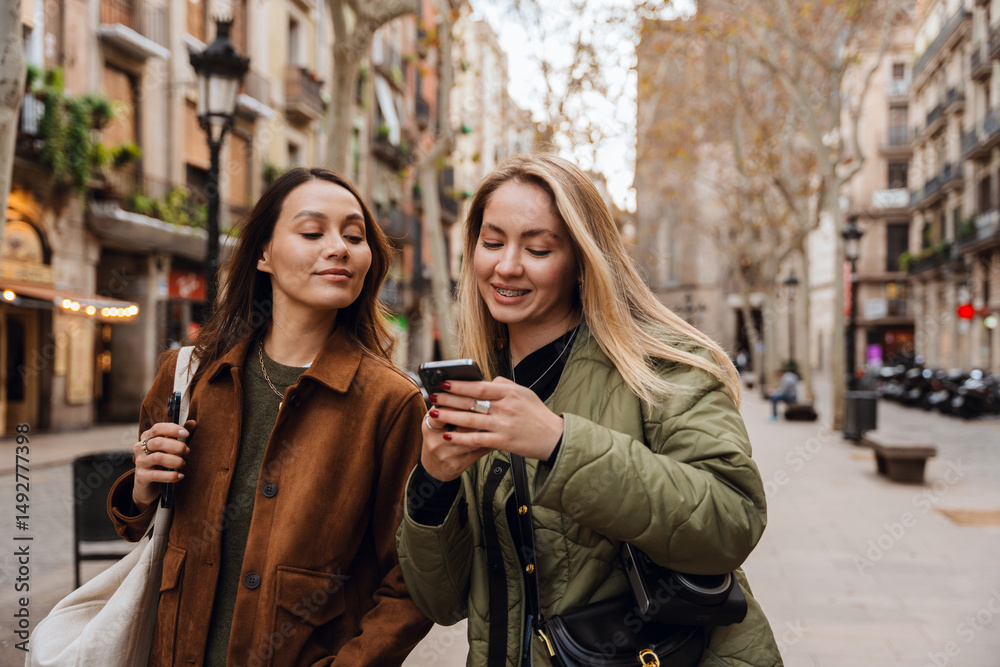 Fototapeta premium Two young White women walk on a city street during the daytime, wearing casual jackets. One woman uses a smartphone while the other looks at the screen.