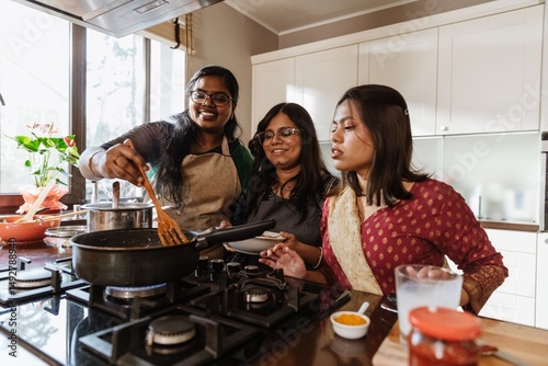 Bild auf Leinwand A group of young Indian women wearing aprons, cooking together in a modern kitchen, stirring a pan, while one woman holds a plate and another adds ingredients, smiling and enjoying the moment