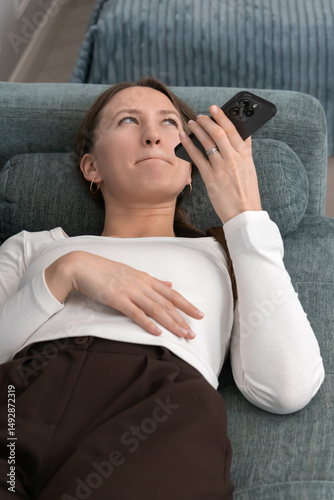 Young woman lying on a cozy sofa, scrolling through her smartphone and expressing annoyance over something encountered online, feeling frustrated and bothered by the content. Vertical photo