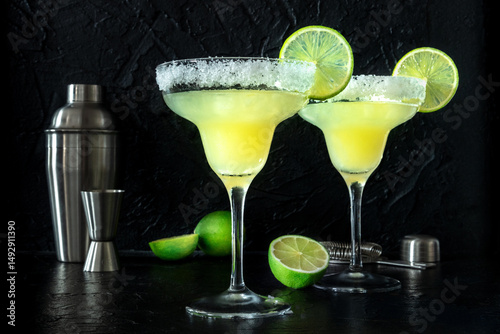 Margarita cocktail in the classic glasses, side view on a black background, with lime slices and a salt rim, with a shaker and a jigger