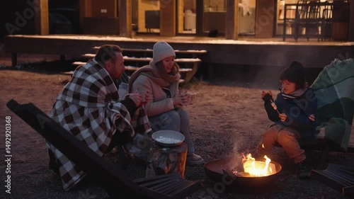A man, a woman, and a school-aged boy sit around a fire pit in the evening near a wooden cabin. The boy roasts marshmallows while the adults drink from cups.
