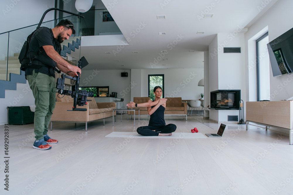 Obraz premium Behind the scene. Photo of a cameraman recording a woman doing yoga exercises at an online training while using and watching a laptop.