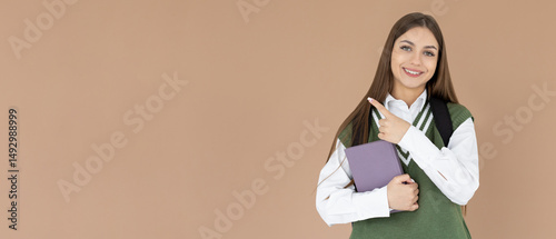 Hey look. Smiling pretty female model pointing fingers right at empty space and inviting to check it out, showing advertisement on brown background. Girl with notepads in hands.
