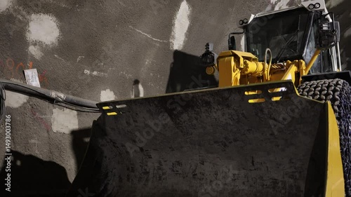 Heavy construction loader closeup with massive bucket and tire chains inside a rough concrete tunnel powerful industrial presence and gritty underground atmosphere dominate the frame with bold intensi