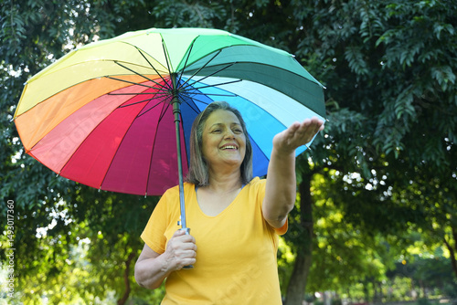 Monsoon Bonanza Concept. Portrait of Indian Senior woman under rainbow umbrella and walking outdoor for relax freedom. Asian elder woman enjoy monsoon with nature, fresh air and stunning views