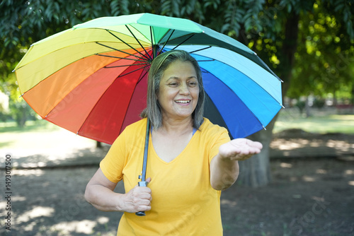 Monsoon Bonanza Concept. Portrait of Indian Senior woman under rainbow umbrella and walking outdoor for relax freedom. Asian elder woman enjoy monsoon with nature, fresh air and stunning views
