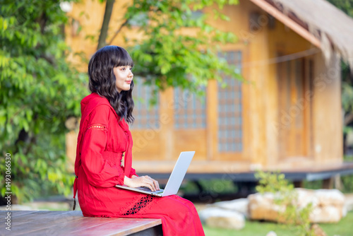 Side view of Asian woman smiling happily while sitting on cottage terrace in the countryside with laptop working remotely on new web design project in sunny garden.