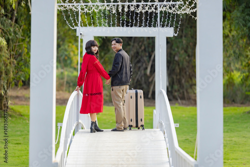 Asian couple with baggage standing with their backs to camera, looking at each other and holding hands against on outdoor quality time together, fun countryside adventure or bonding getaway. 