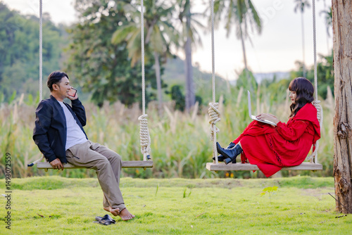 Asian man sitting on swing enjoying mobile conversation, looking at woman using laptop online search for holiday destination or discount offers on travel at beautiful blooming reeds grass flower. 