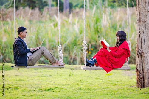 Happy couple enjoying in travel using laptop watching pictures on the laptop and reading book while sitting on the swing at beautiful blooming reeds grass flower blowing from wind.