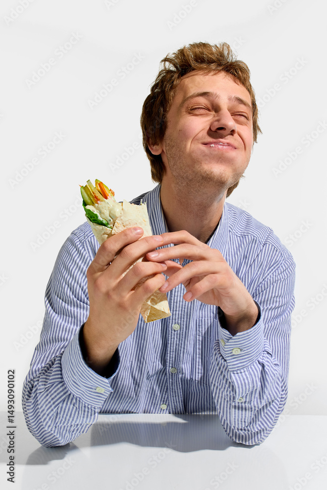 Cheerful man chewing veggie wrap with closed eyes and smiling in striped shirt, hands gently holding tortilla against white background. Concept of traditional, catering, fast food, nutrition, takeaway Cheerful man chewing veggie wrap with closed eyes and smiling in striped shirt, hands gently holding tortilla against white background. Concept of traditional, catering, fast food, nutrition, takeaway