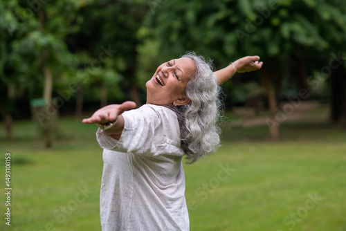 Indian happy senior woman spread her arms in environment nature like the wings of a bird. Happiness. Freedom. Elderly woman stands with one hand open in a park. Indian active old age at woman in park