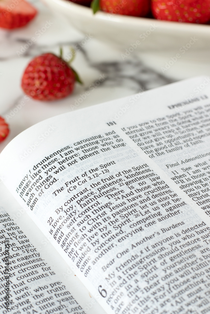 Obraz premium Fruit of the spirit text in open holy bible book with strawberries on table. Biblical concept of love, joy, peace, Christian character growth. Close-up.