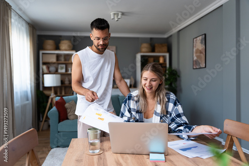 Young couple working remotely at home, analyzing documents and data, using laptop in cozy modern living room, discussing financial reports and business strategy. Couple goals, simple living support.