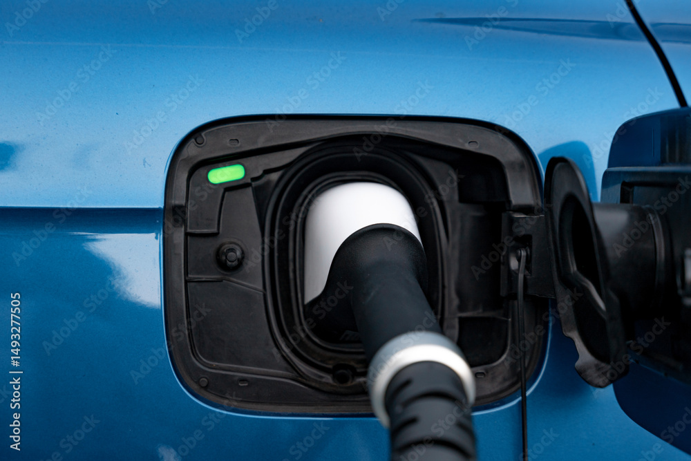 Close-up of Electric vehicle charging station revealing the connector plugged into a vibrant blue car under bright daylight Close-up of Electric vehicle charging station revealing the connector plugged into a vibrant blue car under bright daylight