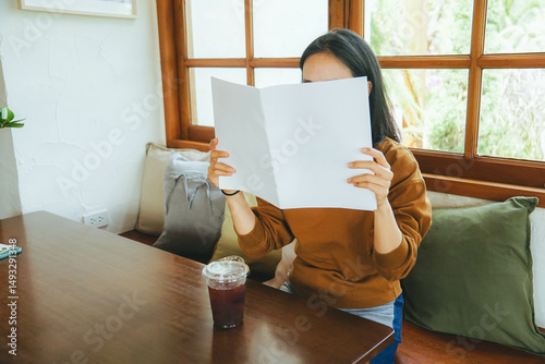 Female covering her face with blank book or magazine and sitting in chair.