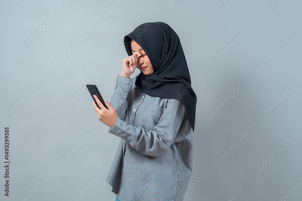 Portrait of a young Muslim woman in a hijab rubbing her tired eyes while holding a smartphone, expressing digital eye strain or screen fatigue in a modern lifestyle context. Portrait of a young Muslim woman in a hijab rubbing her tired eyes while holding a smartphone, expressing digital eye strain or screen fatigue in a modern lifestyle context.