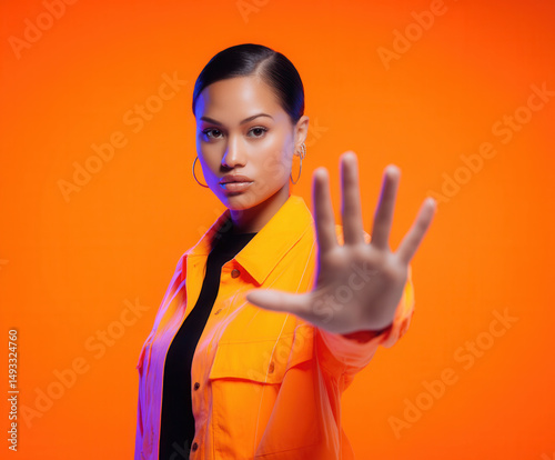 Confident woman gesturing 'stop' against a vibrant orange backdrop, exuding authority and style with her poised demeanor and bold look. photo