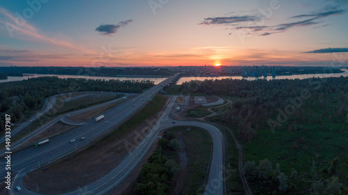 Drone photo of a highway bridge stretching over a river toward a Barnaul city skyline, captured at colorful sunset with clear sky.