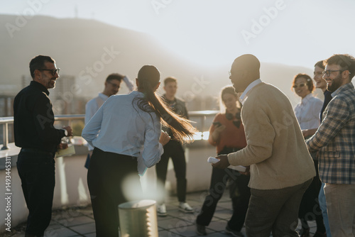 A diverse group of business people celebrate success on a high rooftop at sunset, enjoying music and dancing while sharing joyful moments together against a beautiful cityscape backdrop.