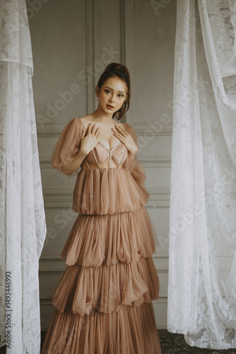 Elegant woman wearing a tiered gown with sheer sleeves poses indoors with curtains