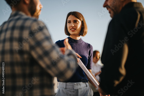 A diverse group of business colleagues engage in a conversation on a high tower balcony during sunset. The scene captures a collaborative and professional atmosphere with a multicultural team.